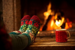 Feet in woollen socks by the Christmas fireplace. Woman relaxes by warm fire with a cup of hot drink and warming up her feet in woollen socks. Close up on feet. Winter and Christmas holidays concept.